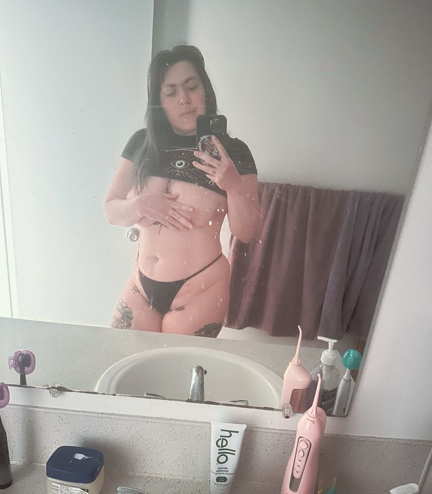 Stormy nightingale is standing taking a selfie in her bathroom mirror. She’s wearing a black thong and black crop top from her illuminaughty burlesque show it’s pulled up to expose her tits that are half covered by her arm