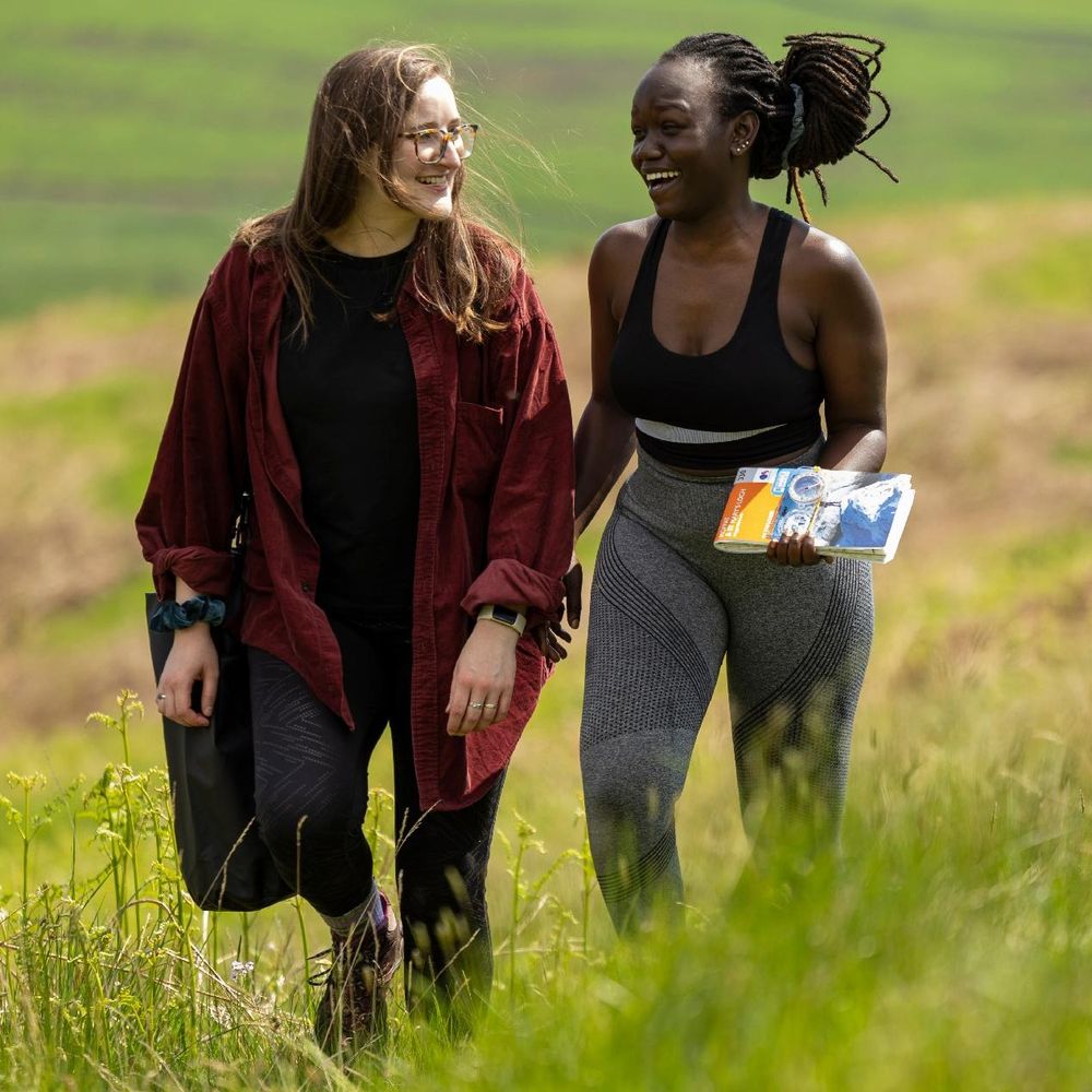 Two people walking through a grassy field, smiling and chatting. One is holding a map.