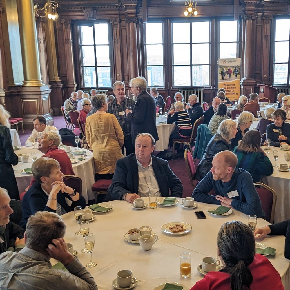 A group of adults gathered in a grand dining hall, engaging in conversation around several banquet tables. Some attendees are seated while others stand, and the room features elegant wooden paneling and large windows.