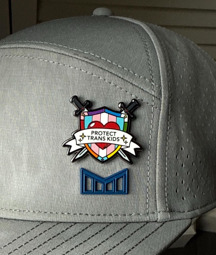 Grey cap with a colorful pin that has the words “Protect Trans Kids” on a white banner over a shield with blue, pink, and white stripes, and crossed swords behind that. Purchased from
Hokum and Snark, a queer-owned Etsy shop.