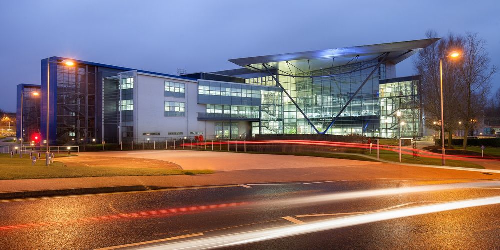 Long exposure picture of Met Office HQ, Exeter at night/twilight. Road in foreground, greenery and path towards glass-plate building behind.