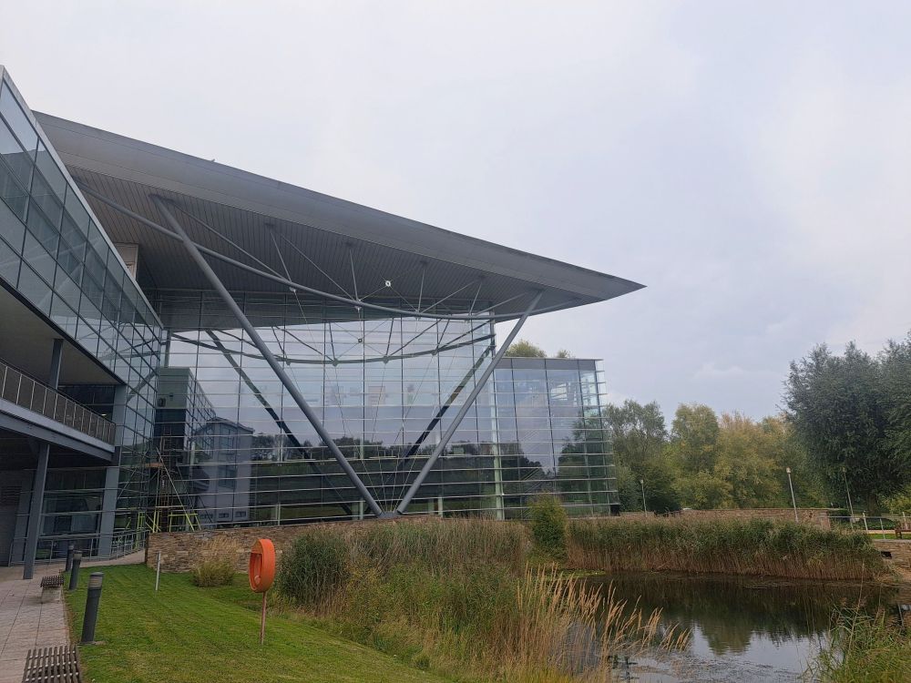 Looking towards Met Office HQ, a glass plated building with a pond and associated greenery in the foreground. Grey sky
