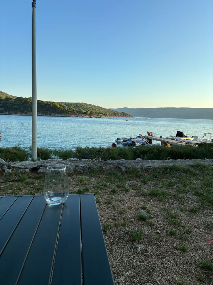 Das Foto zeigt eine friedliche Szene am Meer bei klarem Himmel und Tageslicht. Im Vordergrund ist ein dunkler, rechteckiger Tisch mit einer Glasscheibe zu sehen; darauf steht ein einzelnes, leeres Wasserglas. Der Tisch steht auf einem Erdboden mit etwas Grasbewuchs. Hinter dem Tisch verläuft eine niedrige Steinmauer, dahinter wächst niedriges, buschiges Grün. Weiter hinten erstreckt sich das Wasser ruhig bis zum Horizont. Links am Ufer steht ein hoher, dünner Mast. Auf dem Wasser befindet sich ein kleiner Bootssteg mit mehreren kleinen Booten, rechts davon sitzen oder stehen einige Menschen am Ufer oder auf dem Steg. Am gegenüberliegenden Ufer ist ein bewaldeter Hang zu sehen, der sanft ins Wasser abfällt. Der Himmel ist klar und hellblau, ohne Wolken. Insgesamt wirkt die Szenerie ruhig und einladend, mit der Atmosphäre eines entspannten Sommerabends 