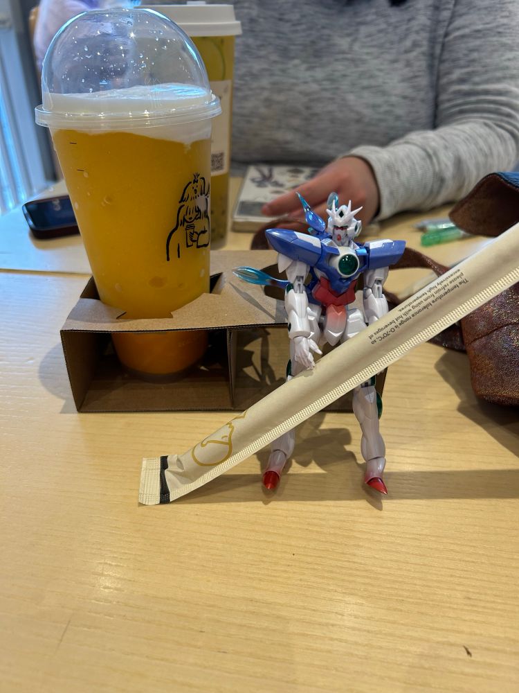 Robot Spirits Side MS ELS Qan[T] holding a drink straw, with a mango slushie behind her.