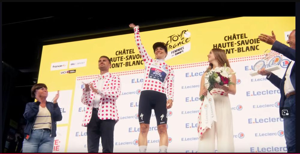 Elise Chabbey’s podium ceremony at the end of the women's Tour - she stands on the top step of the podium in the polka dot jersey of the Queen of the Mountains. To her left (as we're looking at it) is a man in a polka dot suit jacket and bow tie, to her right a woman in a white jumpsuit with a polka dot scarf around her waist holds flowers to give to Chabbey.