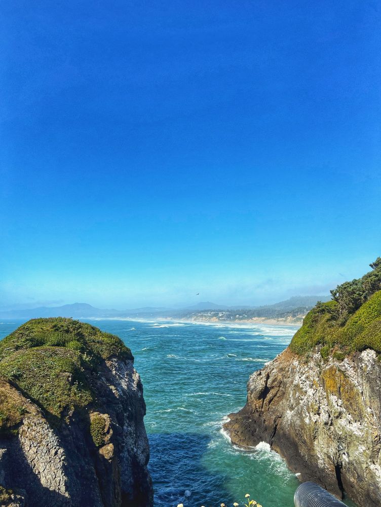 Looking from a cliff that overlooks the Pacific Ocean in Oregon. The ocean is a beautiful blue-green with cliffs and huge rocks in the ocean that have greenery on them. There’s a light fog in the air