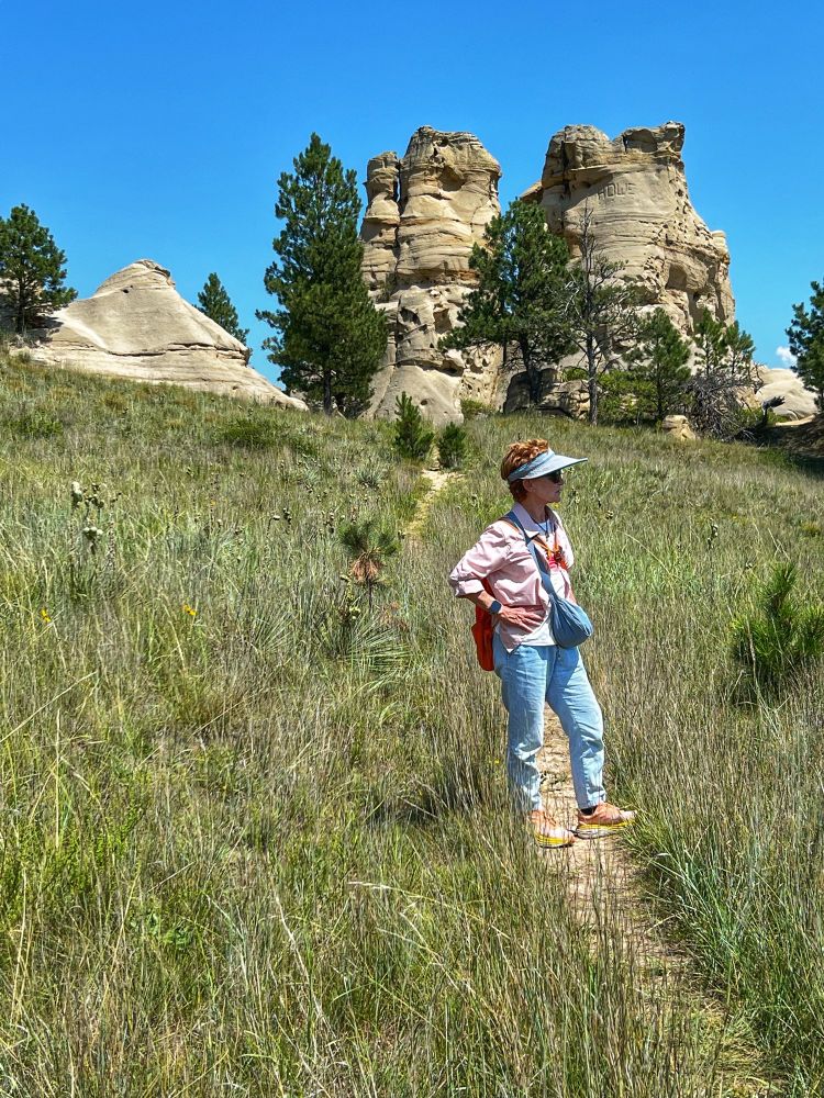 A female, (me! 😁) wearing light blue pants, a peach-colored shirt and a large blue visor on her head is pausing on a hiking path. Behind me are large sandstone rock formations caused by  erosion. 