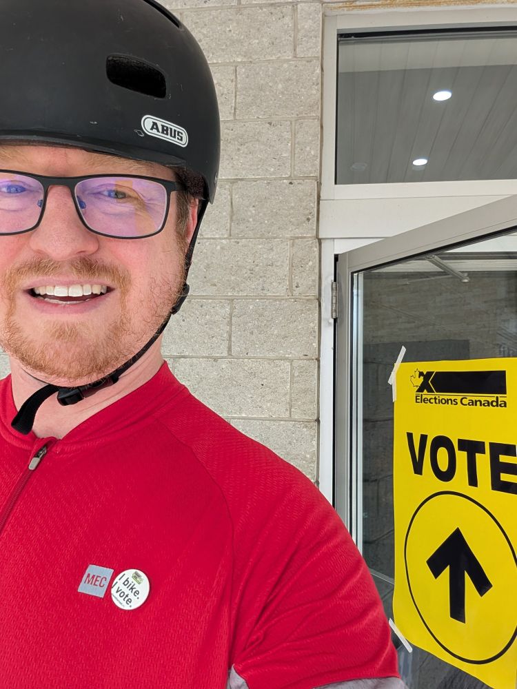 Mike Boos in a red cycling jersey and bike helmet, in front of a polling station. An "I bike, I vote" pin is attached to the cycling jersey.
