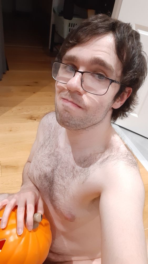 A naked guy with mid length hair stubble and glasses is on the floor hiding their dick with a three tiered pumpkin