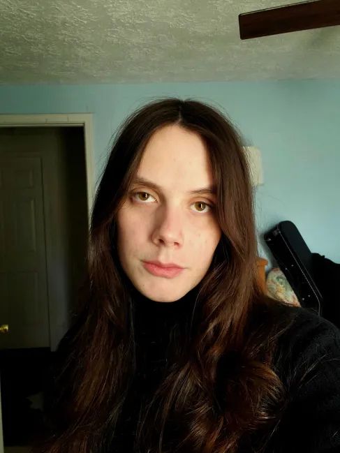 selfie of jo in a black turtleneck, looking into the camera with a half-dead expression. they are softly lit by sun from a window