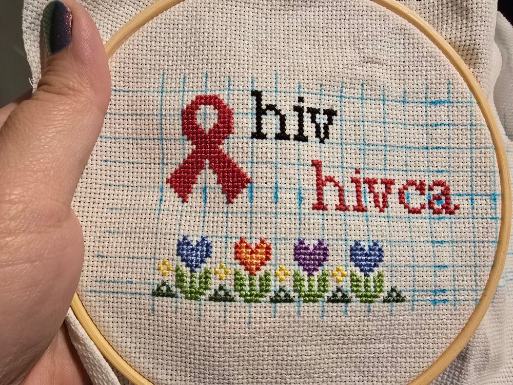 blue grid with white aida. there is a red ribbon on the left-hand side, 'hivca' in red and 'hiv' in black. there are four heart flowers: two with a dark and light blue, one in a red and light red-orange, and a dark and light purple one.