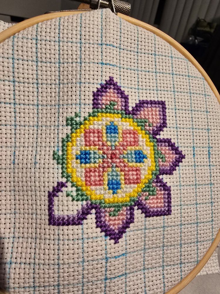 blue grid on white aida. a flower is beginning to form at the center. using yellows. whites, light and dark blues, light and dark purple, and pink.