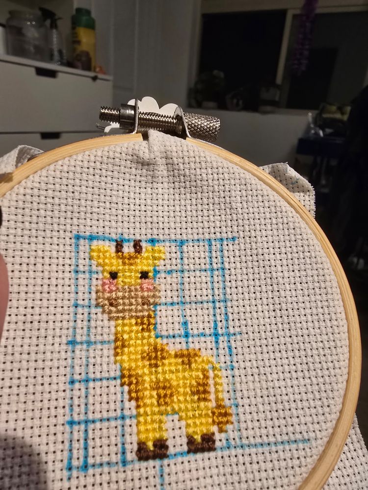 a complete cross stitched kawaii blushing giraffe on white aida. the giraffe is light yellow, golden, coffee brown, and geranium for blush.
