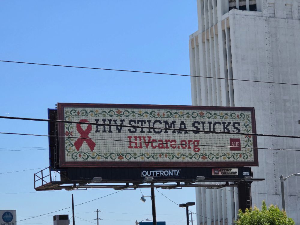 a billboard that says HIV stigma sucks - HIVcare.org. it's printed in a cross stitch style with a floral border, a red ribbon, and has an AHF logo in the bottom right corner.