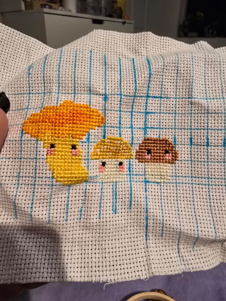 3 mushrooms on white aida with blue grinding. there is a golden chanterelle. gold button, and baby bella with black eyes and a little bit of blush.