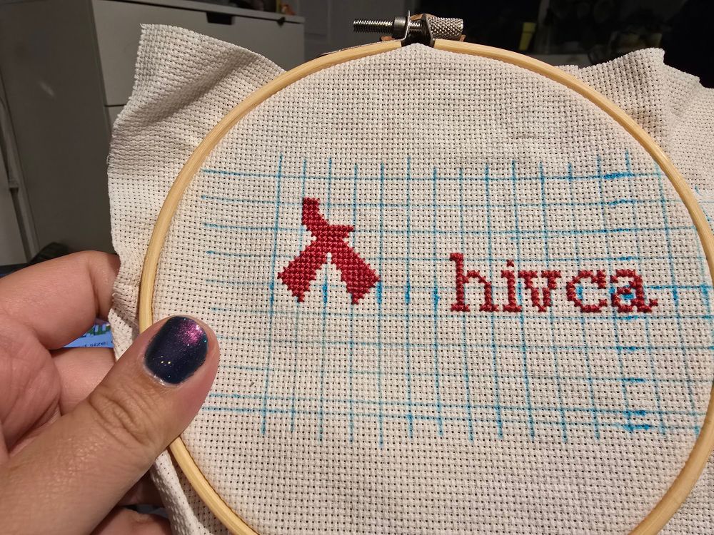 white aida with a blue grid. there are several letter that are starting to form words "hiv ca" and a half-complete red ribbon.