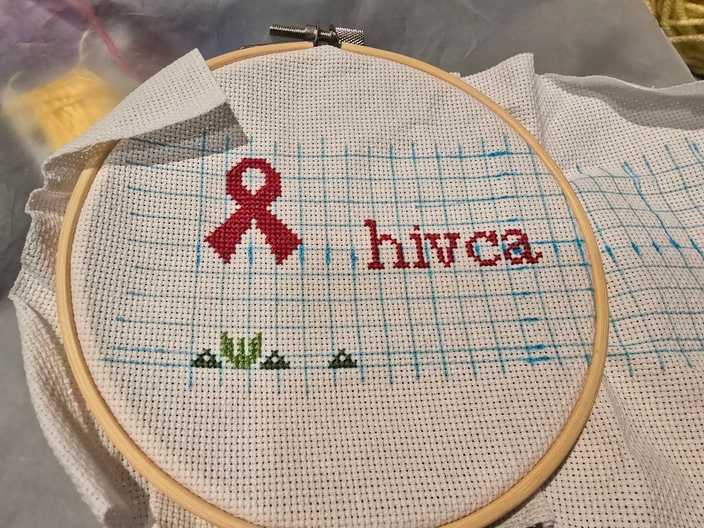 blue grid on white aida. there are some leaves in a light and dark green at the bottom, a red ribbon, and "hivca" to the side.