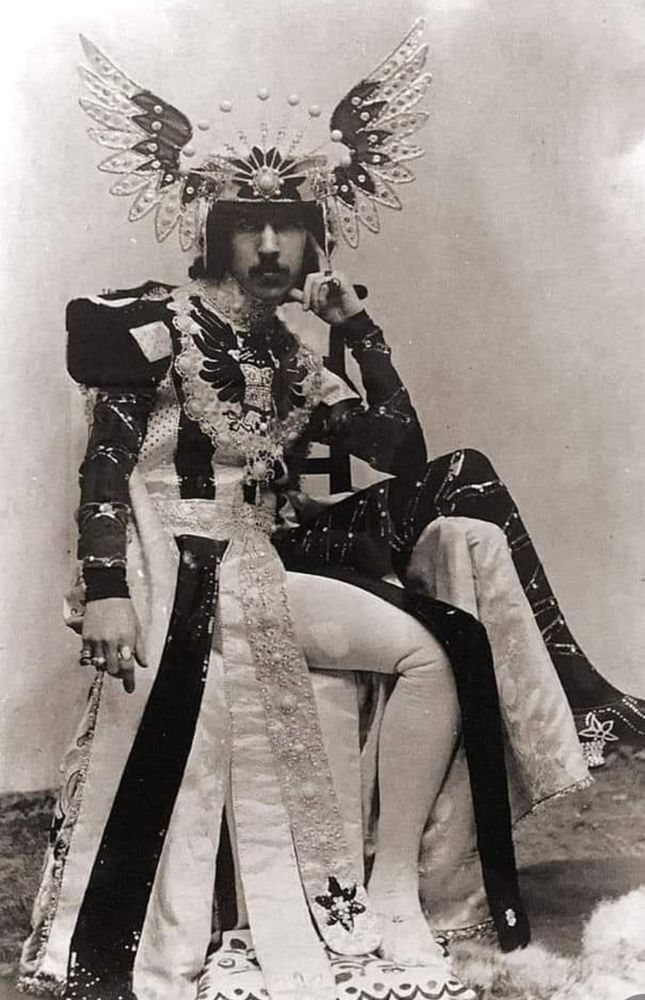 Black and white photo of the Fifth Marquess of Anglesey, sat down with legs to the right and face forward. He is wearing a jewelled and winged helmet and a long robe with the bottom separating into vertical sections. There is a lot of embroidery and adornment. He is resting his left hand on the side of his face.