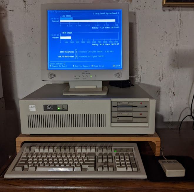 A mid 80s IBM PC/AT with dual 5.25-inch floppy drives running a benchmark program with a brick-shaped Logitech mouse with 3 buttons plugged into it. It matches the color scheme though!