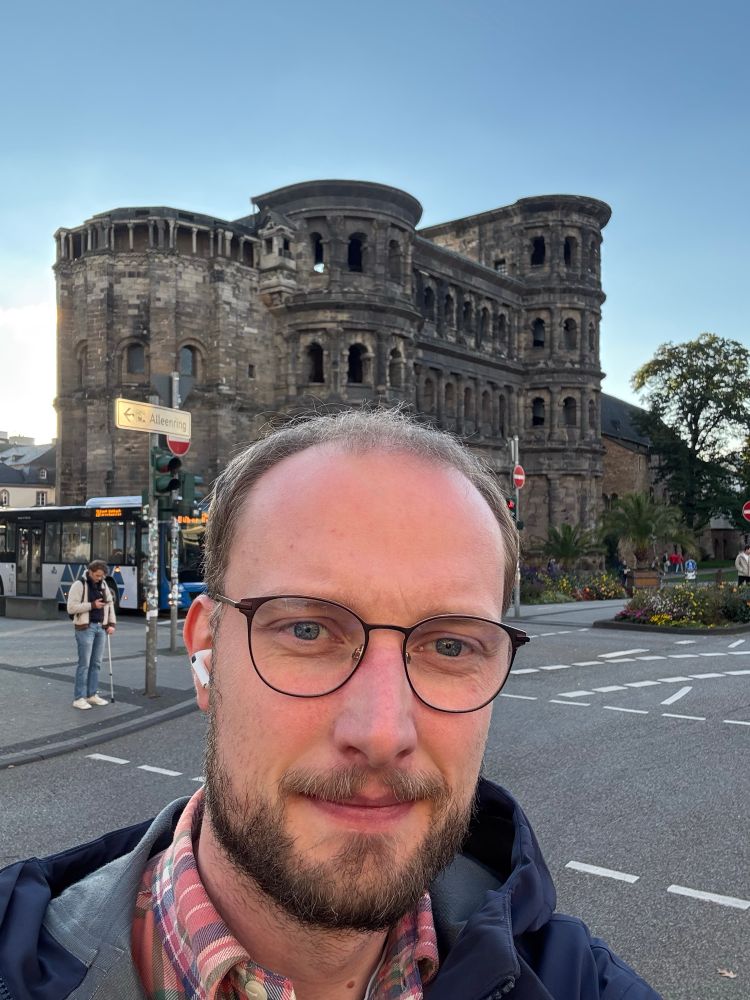 Mann mit Brille (ich) vor der Porta Nigra (stadtabgewandte Seite) in Trier. Es ist gutes Wetter und die Abendsonne erahnbar.