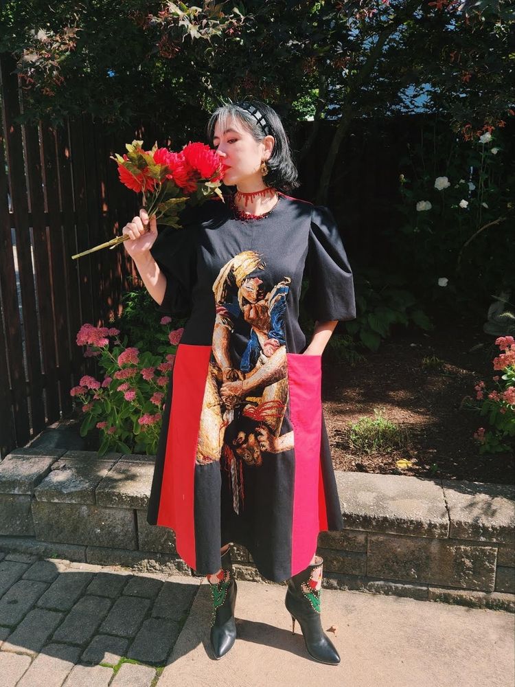Me pretending to smell fake flowers. I’m wearing a loose fitting black dress with red pockets that go down to the hem. In the center is an embroidery and beadwork recreation of Artemisia Gentileschi’s Judith Slaying Holofernes.