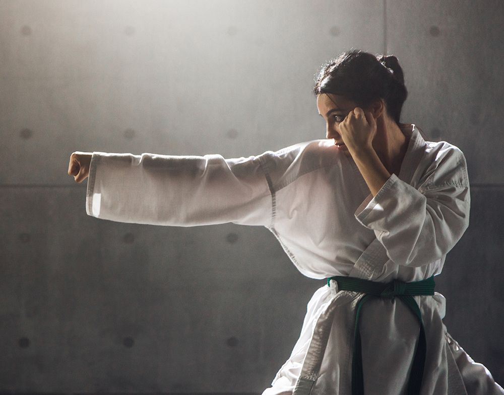A woman in a white karate gi with a green belt strikes a punching stance in profile. One fist is held next to her face and the other fist is extended straight out in front of her. A grey, industrial-looking wall serves as the background.