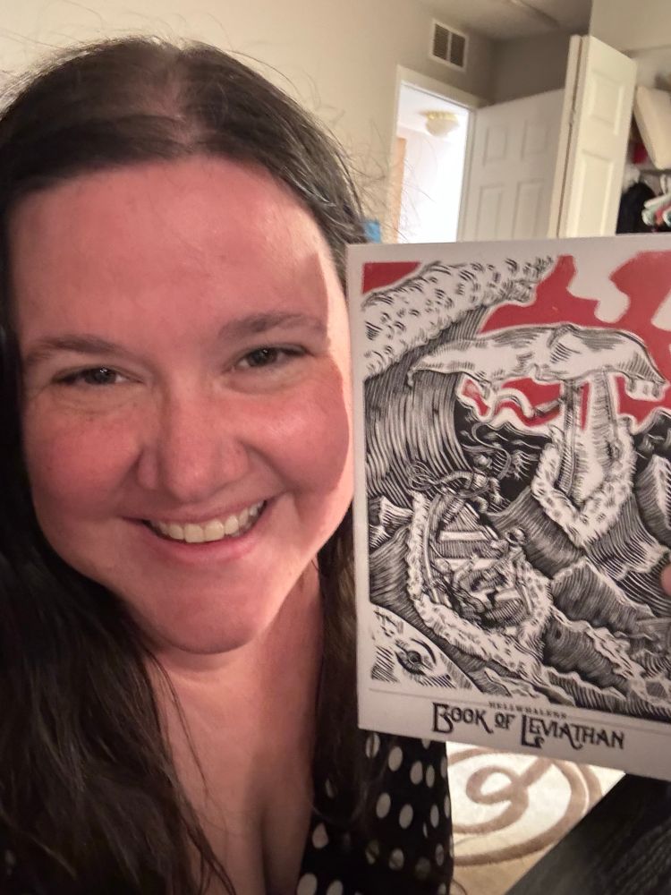 Jocelyn holding up the Book of Leviathan expansion of Hellwhalers, which has a ship on a roiling ocean and a whale.