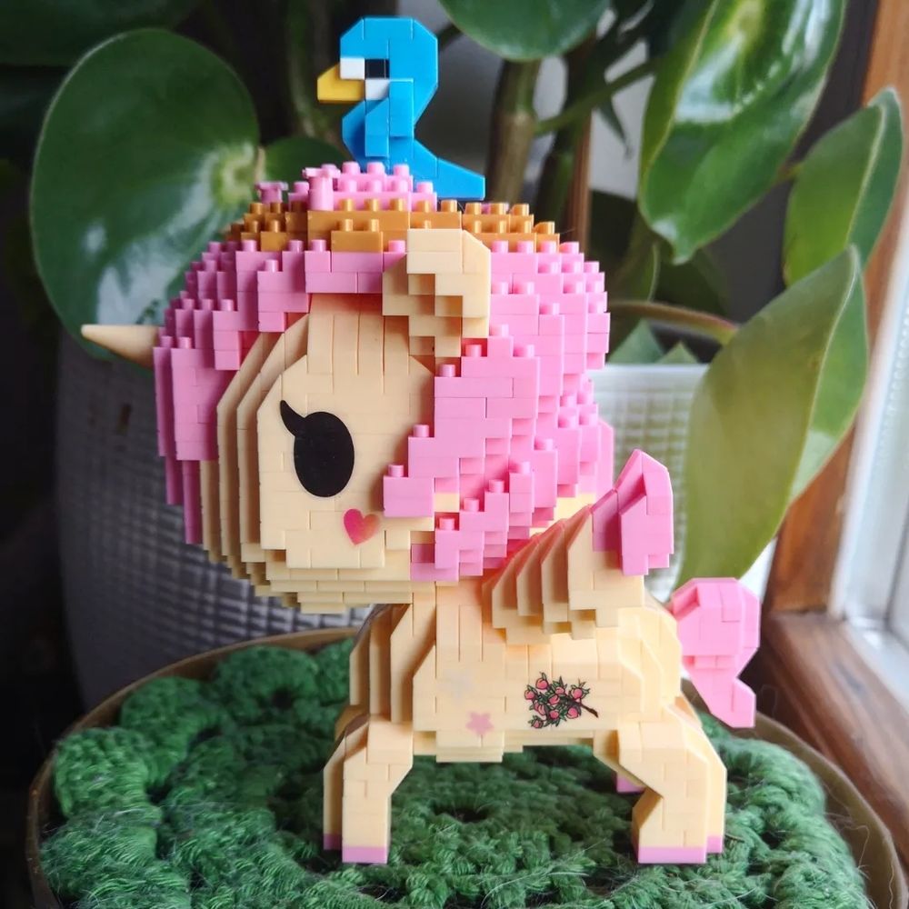 Beigefarbenes Einhorn mit Flügel und rosa Haaren/Schweif aus Klemmbausteinen. 
Auf dem Kopf sitzt ein blauer Vogel.