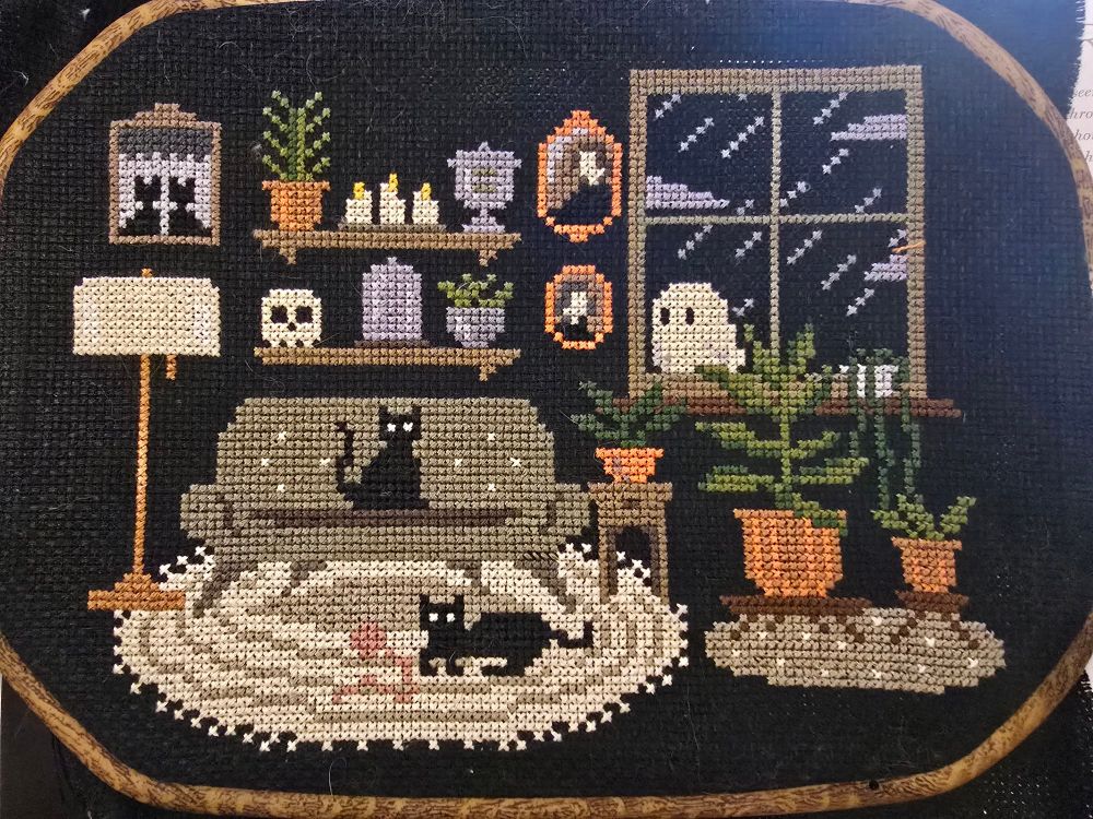 Cross stitch scene of a living room with a couch, a rug, two cata, photos, plants and shelves.