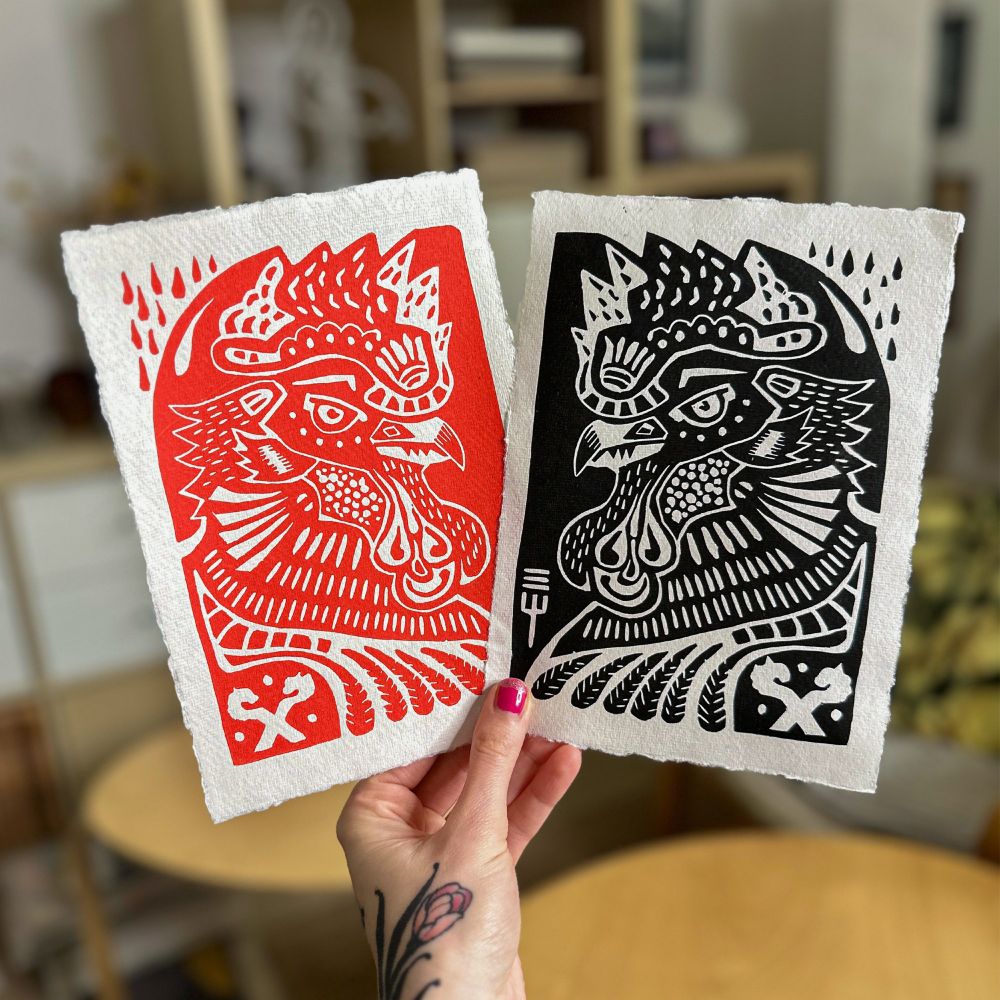 2 linoldrucke in der hand. zwei hähne, einmal in rot, einmal in schwarz. 