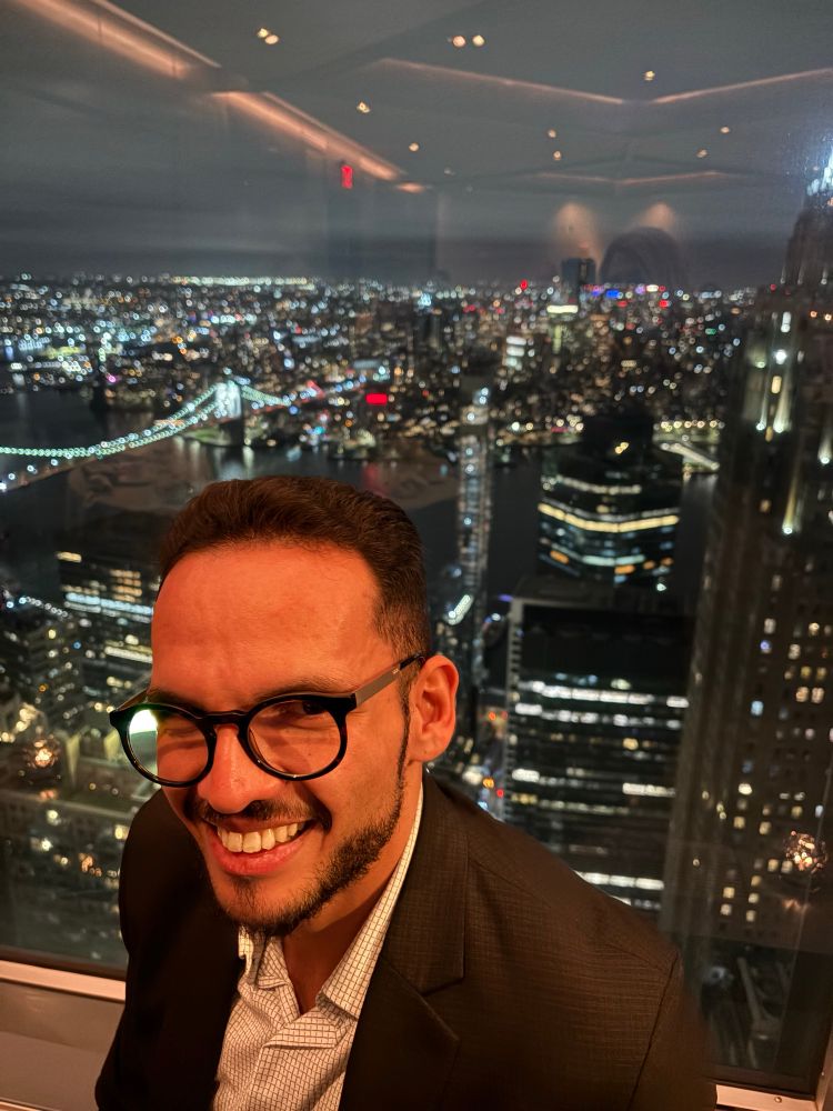 Foto de um gay branco usando óculos com a noite da cidade de Nova York ao fundo 