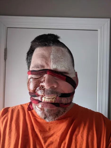 Halloween zombie/frankenstein makeup. Upper right quarter of head is shaved and made paler with makeup. Lower left quarter of face is also shaved but made darker with makeup. Bindings wrap around the head, up against the gums to curl the lips back and permanently expose the teeth, as well as over the eyes to act as a kind of blindfold, save for the corner of one eye to see out of. 