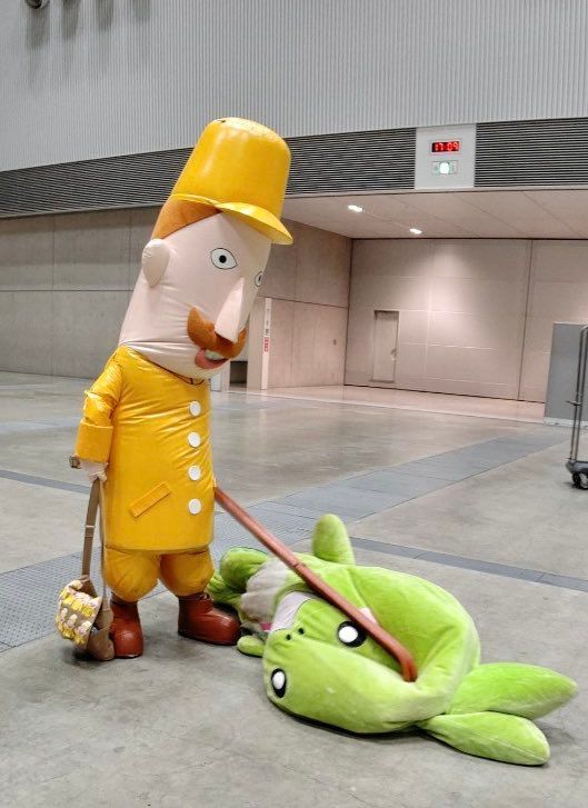 Leruhi-San (left), a tall mascot resembling a cartoon Prussian soldier with brown hair, a curly mustache, goofy smile, and yellow uniform, uses his skis to squeeze the bad juju out of Sasadangon (right, squished on the floor), a round lime green mochi-bamboo-dragon mascot. They’re both mascots from Niigata Prefecture. Leruhi-san represents the Joestu region, famous for its snowy winters and ski resorts, and Sasadangon represents Niigata City. 