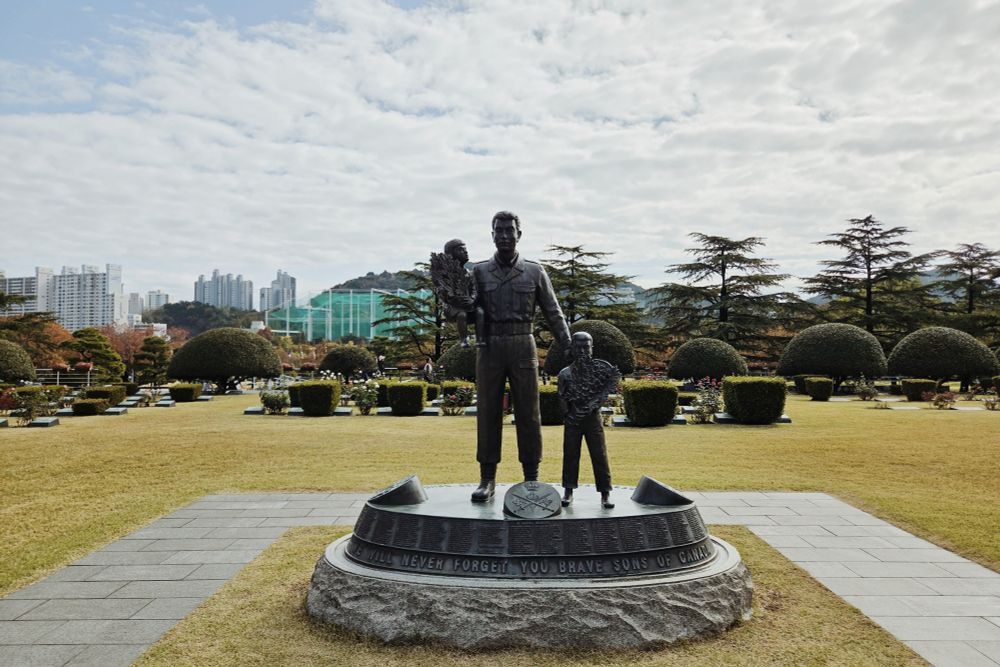 A bronze statue of a soldier with two children holding bouquets. The names of the fallen are inscribed below, their graves, flanked by rose bushes, behind.