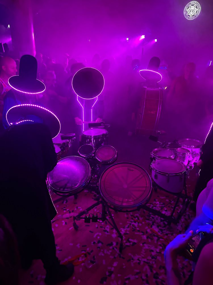 masked figures in black drumming. the figures are wearing large hats with wide brims, the brims are lit with leds. the floor is covered in confetti and there is a haze in the air.