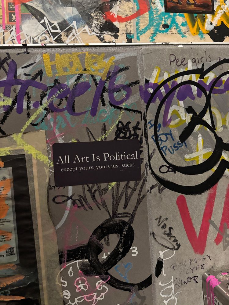 a heavily graffitied bathroom wall. in the center a sticker reads: “all art is political. except yours, yours just sucks.” 