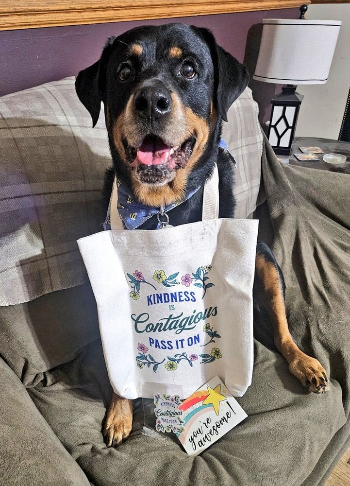 A smiling rottweiler sits on a brown
loveseat with a tote bag around
his neck. There's a screenprint on the bag of pink and yellow flowers
along with the words "Kindness is
Contagious. Pass It On." A sticker with
the same wording and a postcard
proclaiming "You're awesome!" are
leaning against the bottom of the bag.
