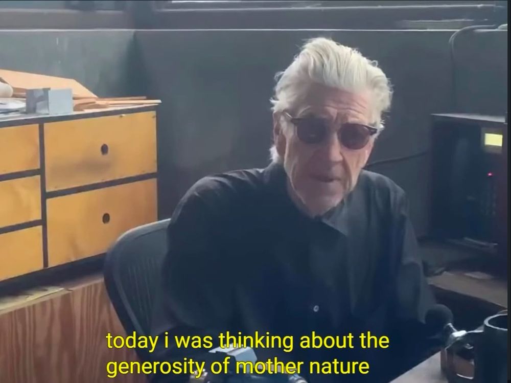 Screenshot from a video or livestream of David Lynch. He’s sitting at his desk in a black dress shirt buttoned all the way up. He has sunglasses on but is clearly looking at the screen. The caption says “today i was thinking about the generosity of mother nature.”