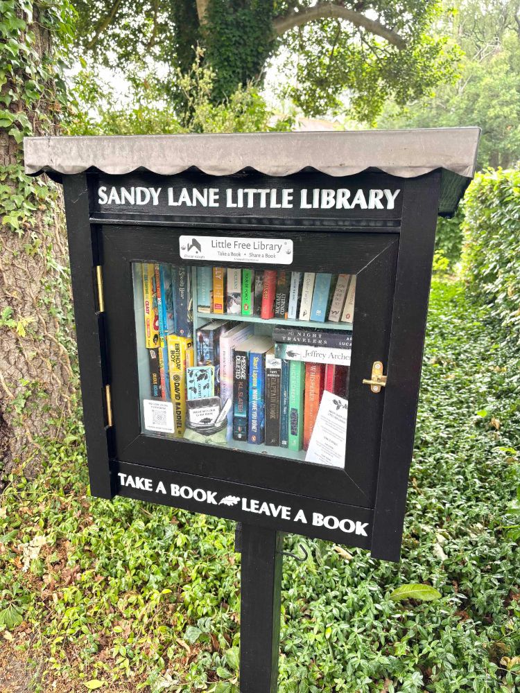 A small black box on a post with glass-panel latched door. Inside is a small selection of mainly paperback books.The box is labelled "Sandy Lane Little Library" and "Take a book, leave a book". A homeowner's contribution to the neighbourhood avid readers!