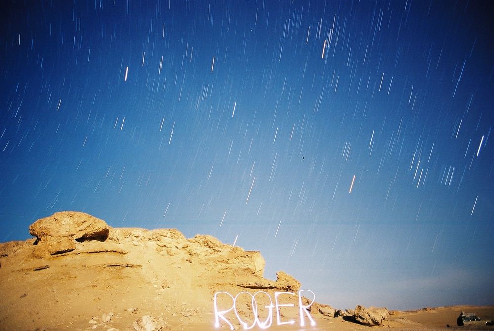 A raw, unprocessed photograph taken at night with a camera on a tripod with c. 15 mins time lapse showing short star trails. The name Roger has been written using a head torch partly revealed to the camera as it traversed the path of each of the letters of the name.