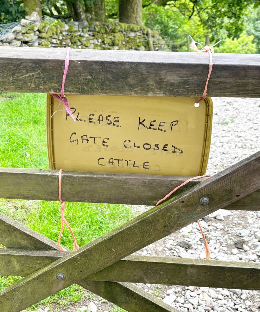 A wooden cross-slatted gate with stone path and grass beyond. A hand-written sign reads "Please Keep Gate Closed" The word "Cattle" is written below.