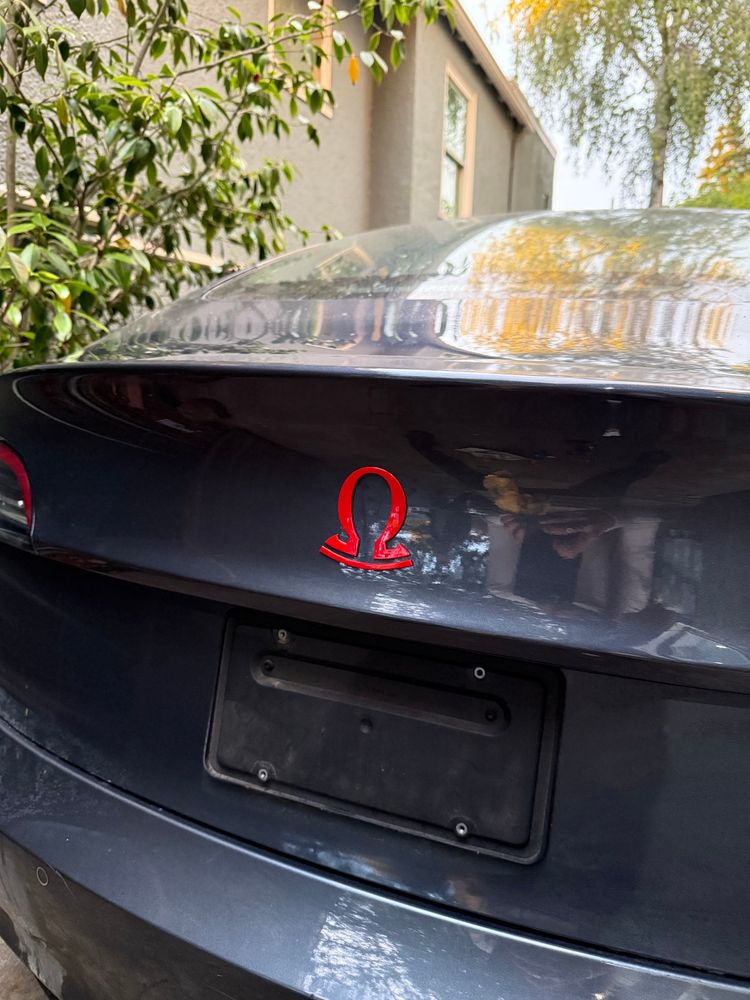 The read of a dark gray electric vehicle. The Tesla badge has been replaced by a stylistically similar red Omega badge.
