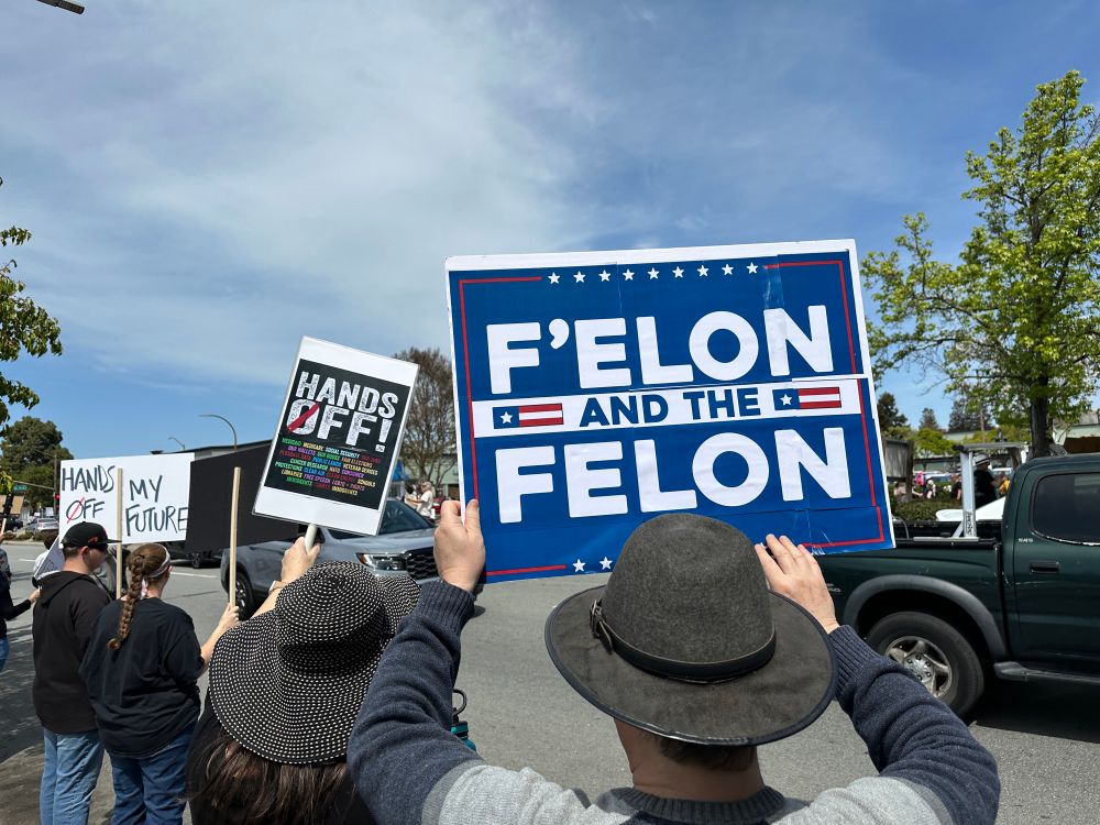 Crowd signs that say “F Elon and the Felon”, and “Hands Off my Future” waving to the cars driving past