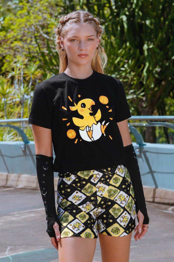 Image of a blonde model wearing a short sleeve black shirt with a super cute chocobo chick bursting out of its shell with an excited expression. Model is wearing high waisted chocobo short shorts as well.