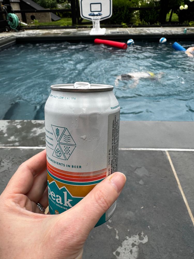 My hand holding a can of slim hazy. A pool is in the background with my kid swimming in it. 