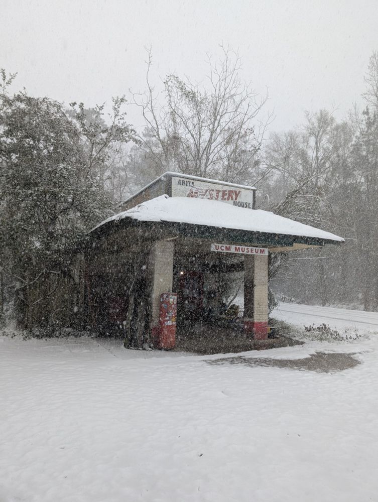 A tiny old-timey corner gas station building with hand painted signs that read "Abita Mystery House" and "UCM Museum". Some old tools and signs sit in the covered space and trees are visible behind the building, but everything is covered with a soft layer of snow several inches thick, and more flakes are falling in front of the camera