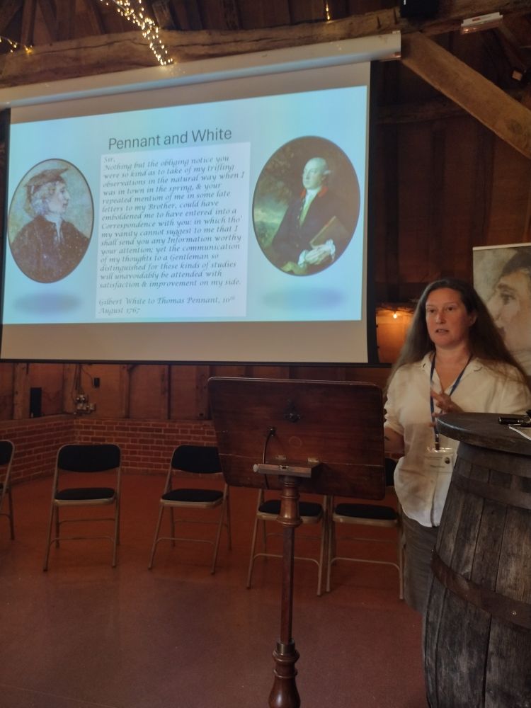 A woman with long hair in a white shirt speaks at a wooden lectern