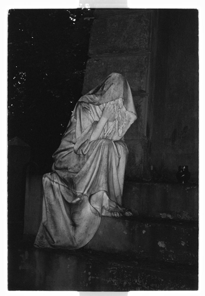 “She weeps, the crypt listens, the lamp stays silent.” (Kodak Professional Plus-X 125)
2022.08.11

Grief Frozen on the Step.
Vvedenskoye Cemetery. Tomb of the Fuld family. A marble mourner sits by the crypt, as if waiting for time to start moving again.
__
Among Lutheran graves, a marble mourner sits — the tomb of merchant Robert Fuld’s family. Her posture embodies grief, her veil breathes through stone. Created in the 1920s, the monument became a symbol of memory that outlasted eras and decay.
__
The mourner sleeps, the lamp never burns out — time does not move.

Technical Credits: 
Location: Vvedenskoye Cemetery, Moscow, Russia
Camera: Samsung Vega 77i QD
Film: Kodak Professional Plus-X 125 (expired December 2012)
Development: Kodak D-76
Scanner: Fujifilm Frontier SP-3000 (no corrections applied — raw scan)
Date: August 13, 2022
Notes: Film stored under unknown conditions for ~10 years past expiry. Results are experimental — a dialogue between time, chemistry, and chance. Grain softened, contrast reduced, tonal range subtly shifted. Each frame carries the memory of its own decay. 