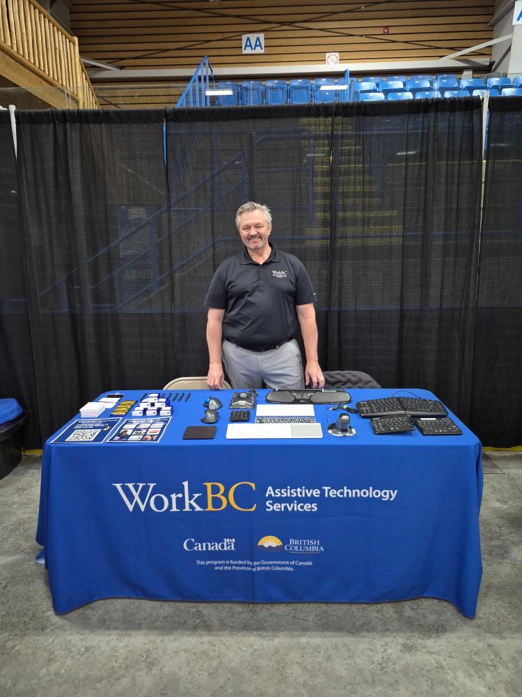 Nate standing at the WorkBC Assistive Technology Services table with assistive technology on display.