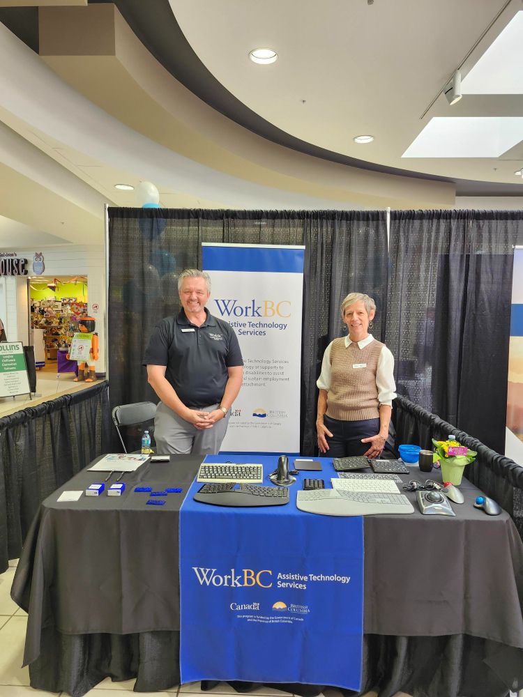 Nate and Tina standing at the WorkBC Assistive Technology Services table with assistive technology on display.